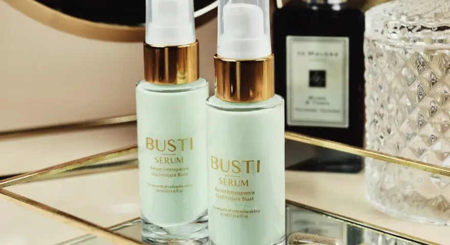 Dwa flakoniki serum ujędrniającego do biustu Busti, kapsułki i eleganckie flakony w tle.