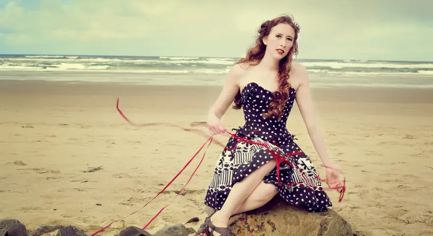 Jeune femme au style pin-up année 50, assise sur un rocher sur la plage, robe à pois, ruban rouge flottant au vent.