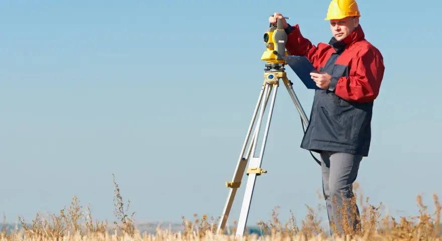 Geodeta w żółtym kasku pracuje z tachimetrem w polu. Zastanawiasz się, ile geodeta bierze od punktu?