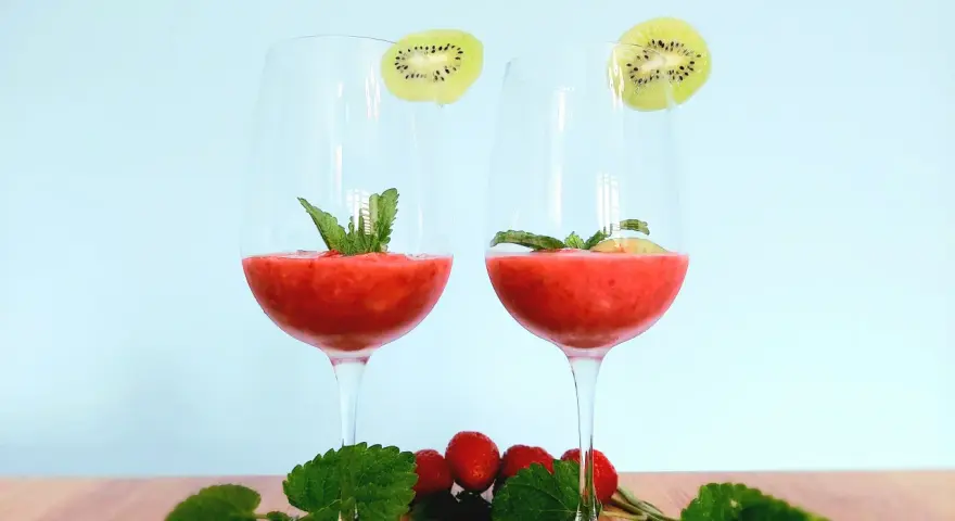 Dwa kieliszki orzeźwiającego drinka truskawkowego z rumem, ozdobione plasterkami kiwi i miętą. Na drewnianym blacie leżą świeże truskawki i listki mięty.