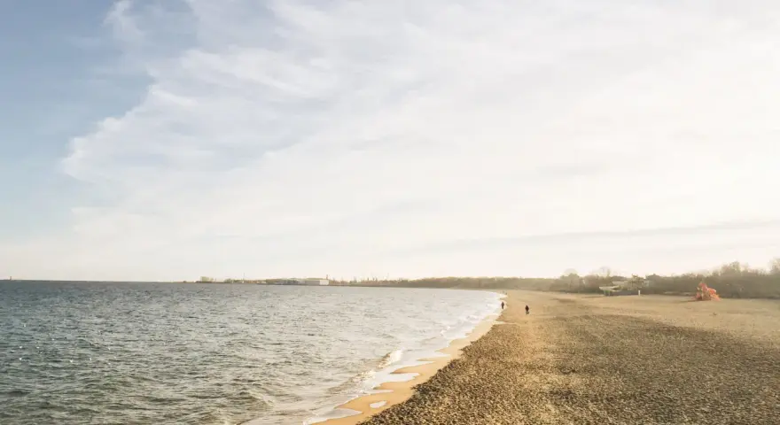 Spokojny brzeg jeziora Białe Kaszuby, fale delikatnie muskają piaszczysty brzeg, a na horyzoncie widać zarys zabudowań.