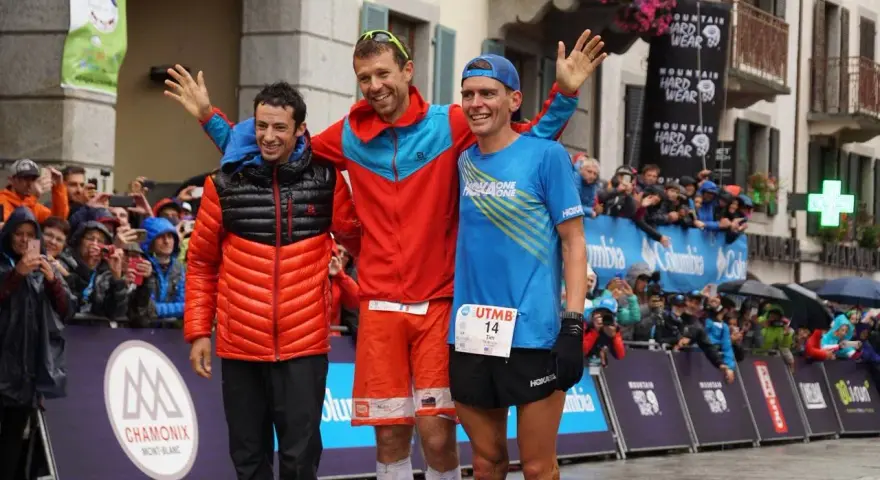 Trzech biegaczy na podium po ultra-trail du mont-blanc. Jeden w pomarańczowej kurtce, drugi w czerwonej, trzeci w niebieskiej koszulce.