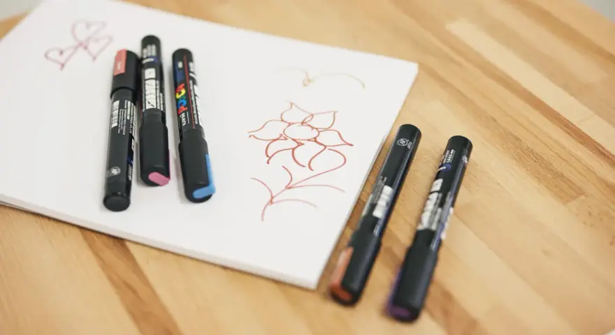 Några Posca-pennor ligger på ett vitt papper med en enkel blomma och hjärtan ritade i rött.
