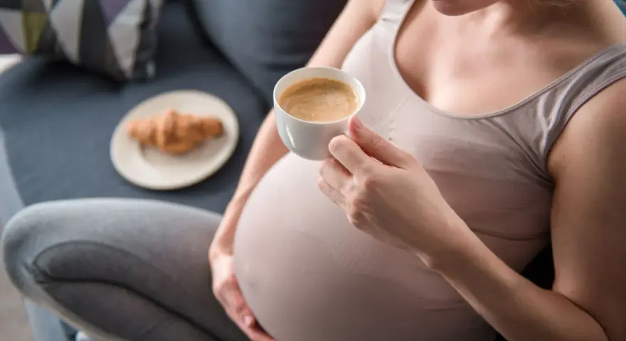 Kawa w ciąży, 1 trymestr. Kobieta w ciąży pije kawę, obok leży rogalik.
