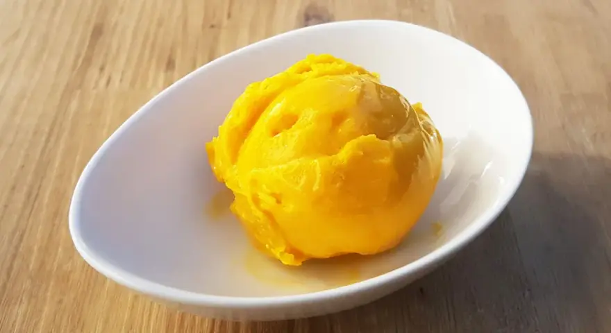 Ein leuchtend gelber Löffel Mango Sorbet in einer weißen Schale auf Holzhintergrund.