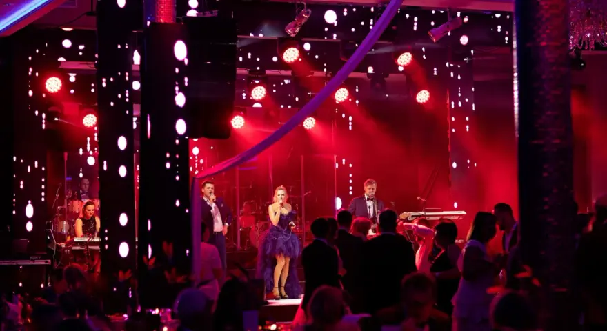 Sylwester w Hotelu Gołębiewski: zespół na scenie, wokalistka w fioletowej sukni, muzycy i publiczność bawią się przy czerwonych światłach.