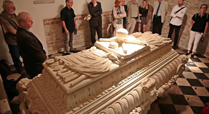Grupa zwiedzających ogląda kamienny sarkofag w wawel krypty. Kamienne rzeźbienia zdobią jego bok.