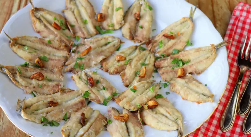 Un plato blanco con boquerones fritos, dorados y crujientes, espolvoreados con perejil fresco y trocitos de ajo.