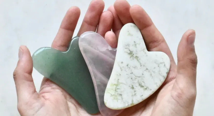 Drei herzförmige Gua Sha Steine in Grün, Rosa und Weiß liegen in einer Hand. Ideal für Gua Sha gegen Falten.