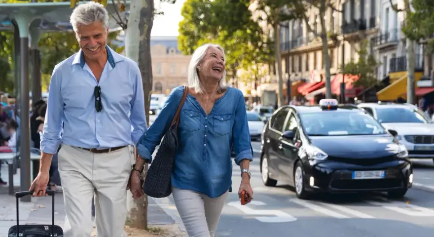 Uśmiechnięta para seniorów spaceruje po mieście z walizką, obok przejeżdżają samochody.