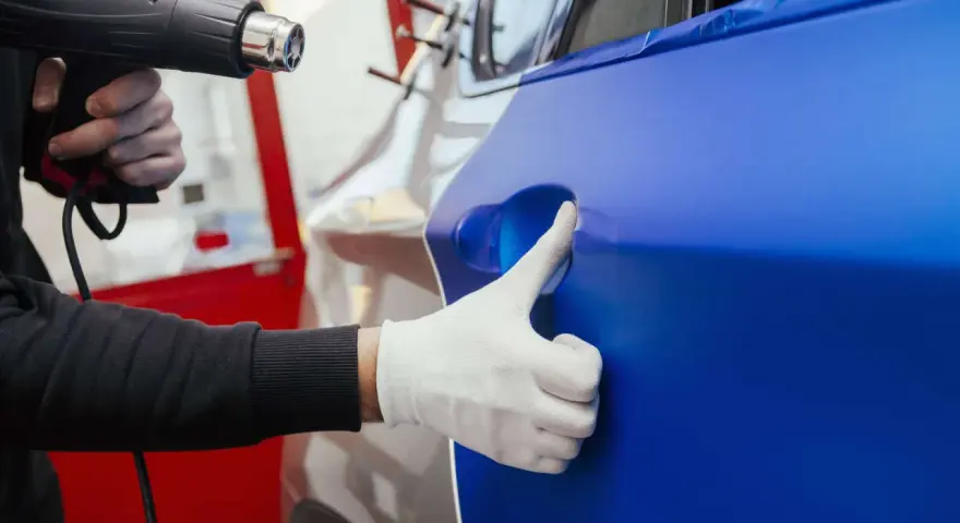 Ein Handwerker erwärmt mit einem Heißluftföhn eine blaue Folie, die auf ein Auto aufgebracht wird. Die auto folieren kosten sind hierbei ein wichtiger Faktor.