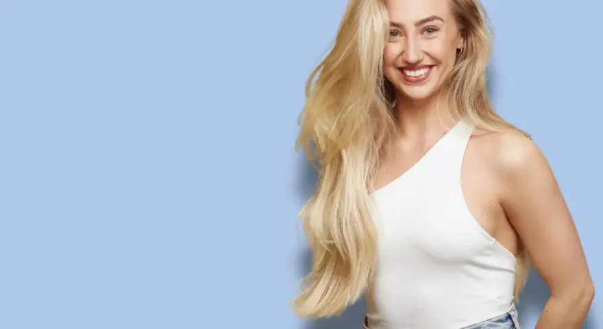 En leende kvinna med långt, blont hår som ser ut att bleka håret naturligt. Hon bär en vit topp och jeans mot en ljusblå bakgrund.