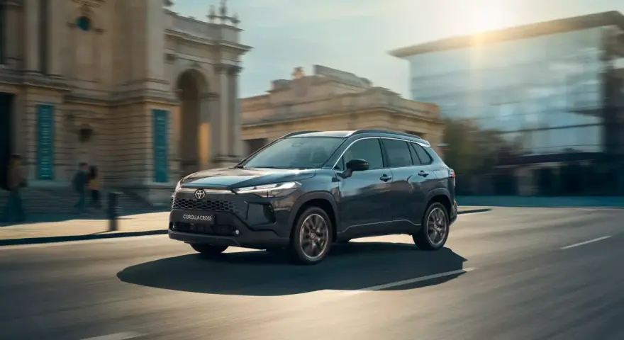 Ciemnoszary Toyota Corolla Cross Hybrid sunie ulicą miasta, odbijając światło słoneczne od nowoczesnych budynków.
