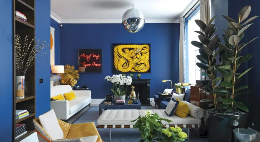 Salón moderno con paredes azul cobalto. Sofá blanco, sillón mostaza y cojines amarillos. Arte abstracto y plantas. Colores que combinan con el azul resaltan.