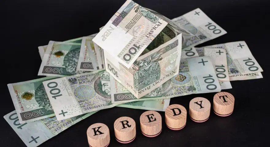 Dom z banknotów 100 zł symbolizuje marzenia o własnym lokum. Drewniane pieczątki z literami K, R, E, D, Y, T układają się w słowo "KREDYT", sugerując potrzebę finansowania lub **restrukturyzacja kredytu**.