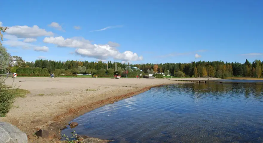 Solig strand vid Nydala camping Umeå med klart vatten och grönskande skog i bakgrunden.