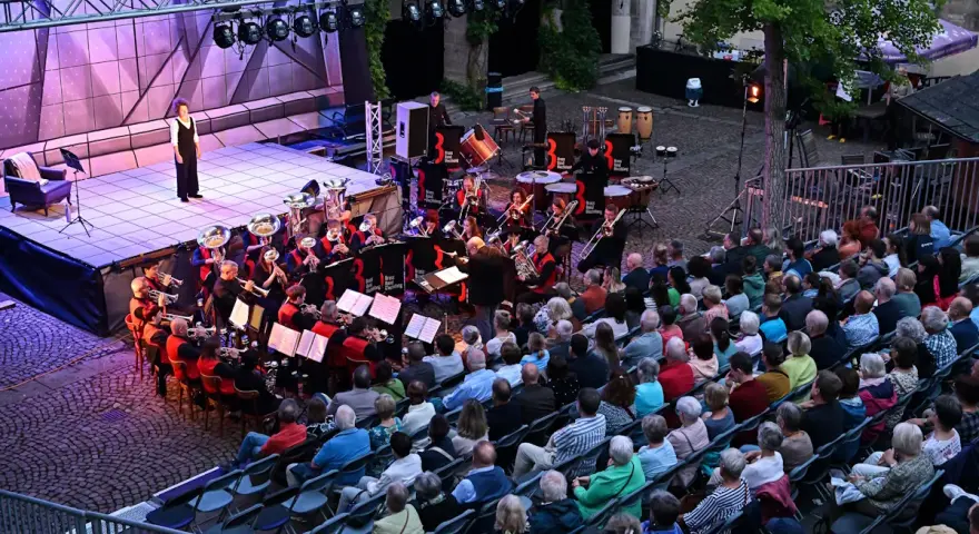 Eine Brass Band spielt auf einer Bühne, während ein Publikum auf Stühlen sitzt und zuhört.