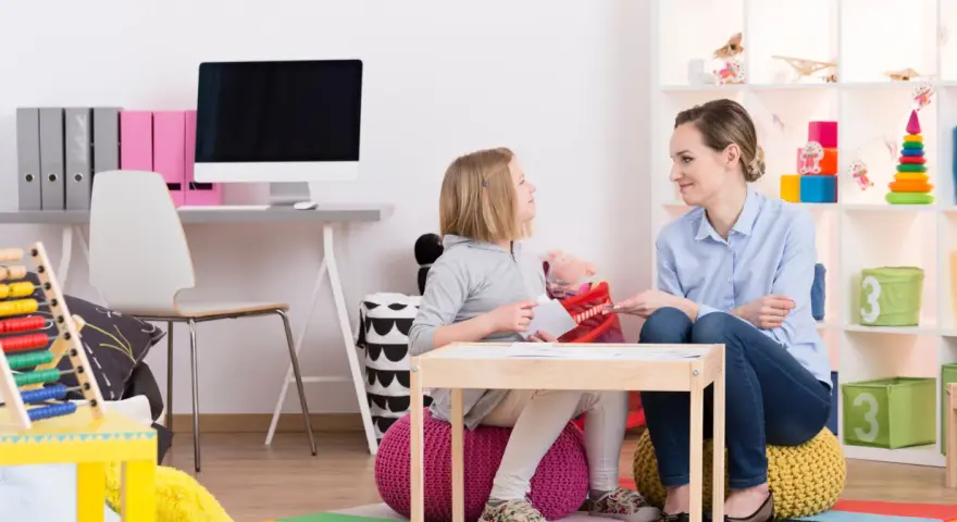 Terapeutka pracuje z dziewczynką, wspierając ją w terapii afazji u dzieci. W tle widać pomoce dydaktyczne i zabawki.