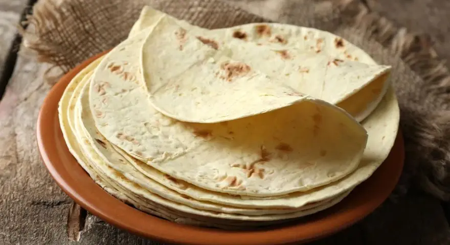 Stos cienkich, jasnych placków keto tortilla na brązowym talerzu, ułożonych na płótnie i drewnianym blacie.