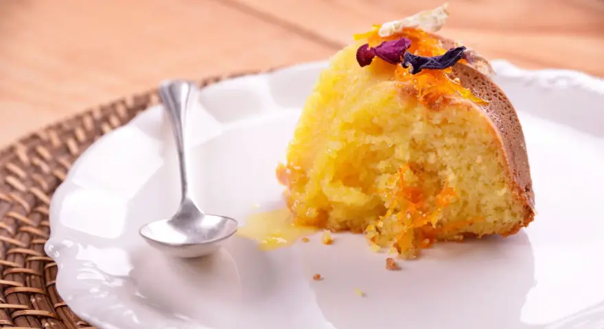 Un trozo de bizcocho de almendra de la abuela, decorado con ralladura de naranja y flores secas, servido en un plato blanco con una cucharilla.