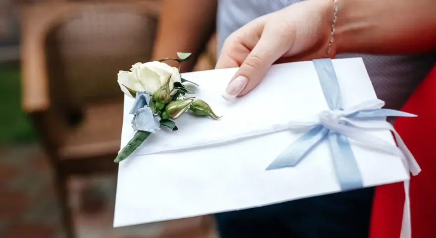 Dłoń trzyma białą kopertę z błękitną wstążką i ozdobnym kwiatkiem. Rozważasz skutki wręczenia pustej koperty lub zbyt małej kwoty? Opinie i etyka są tu kluczowe.
