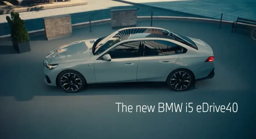 Jasnoszary sedan BMW i5 eDrive40 stoi na zewnątrz, z widokiem na basen i ławkę.