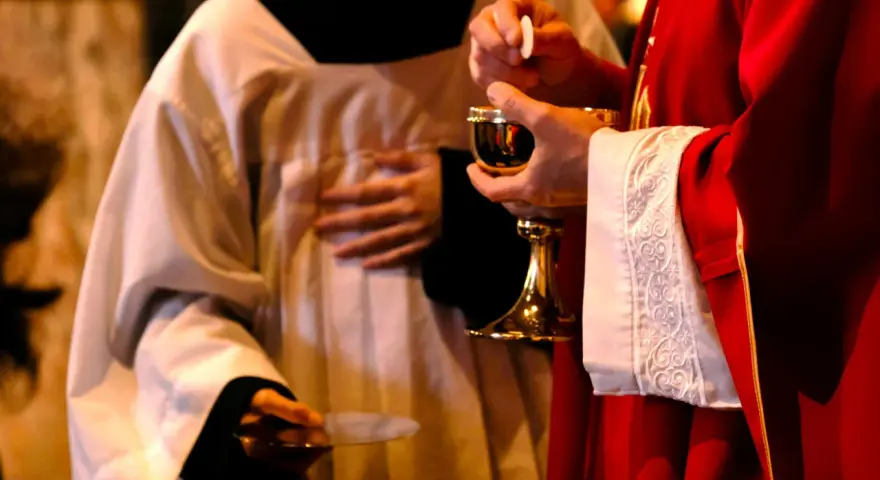 A priest in red vestments holds a chalice and host, while another in white vestments holds a paten. A moment of communion, perhaps a prayer to St. Benedict for healing.