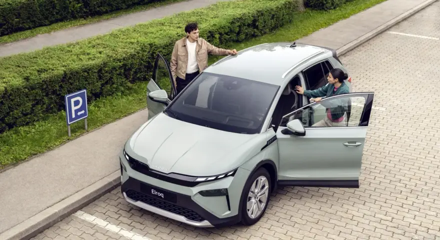 Jasnoszary elektryczny SUV Škoda Elroq z otwartymi drzwiami, obok którego stoją młoda kobieta i mężczyzna.