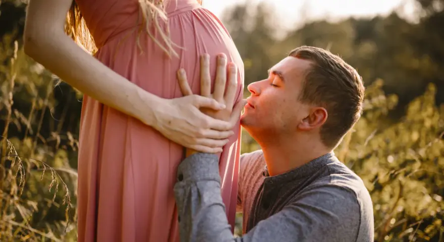 Ciepła sesja ciążowa z mężem. On klęczy i całuje brzuszek ukochanej, otoczeni złotym światłem i naturą.