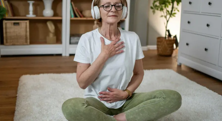 Kvinna med hörlurar sitter på en matta och utövar meditation och andning. Hennes ögon är slutna och hon har händerna på bröstet och magen.