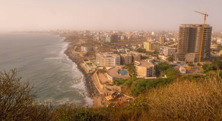 Widok na wybrzeże i rozwijające się miasto w **Afryce, Senegal**. Widać ocean, plażę, nowoczesne budynki i plac budowy z dźwigiem.