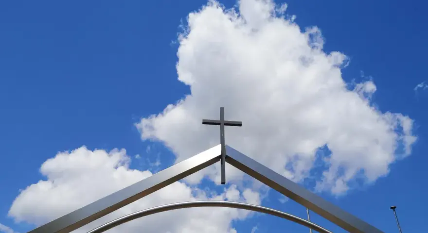 Ein Kreuz ragt über einem modernen Torbogen in den blauen Himmel mit Wolken. Ein starkes Christi Himmelfahrt Symbol.