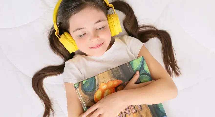Dziewczynka w żółtych słuchawkach leży z zamkniętymi oczami, przytulając książkę. Słucha bajek, które są świetnymi podcastami dla dzieci.