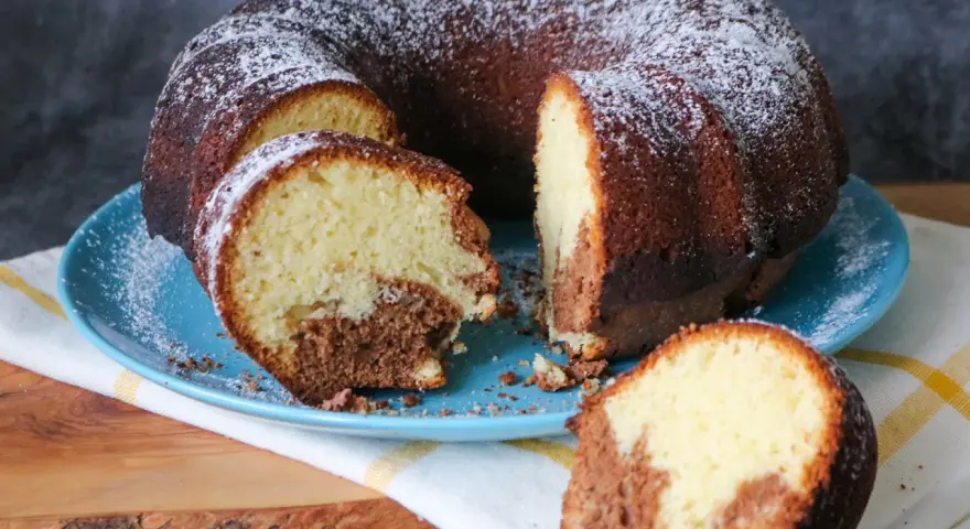 Saftiger weltbester Marmorkuchen, mit Puderzucker bestäubt, auf blauem Teller. Ein Stück ist abgetrennt und zeigt die köstliche Marmorierung.
