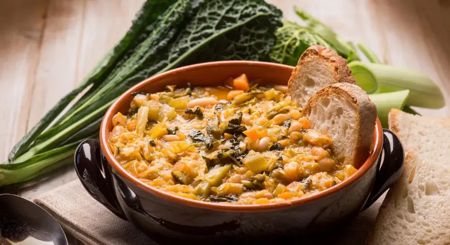 Una ciotola di ribollita fumante, ricca di verdure e fagioli, accompagnata da fette di pane tostato e cavolo nero.