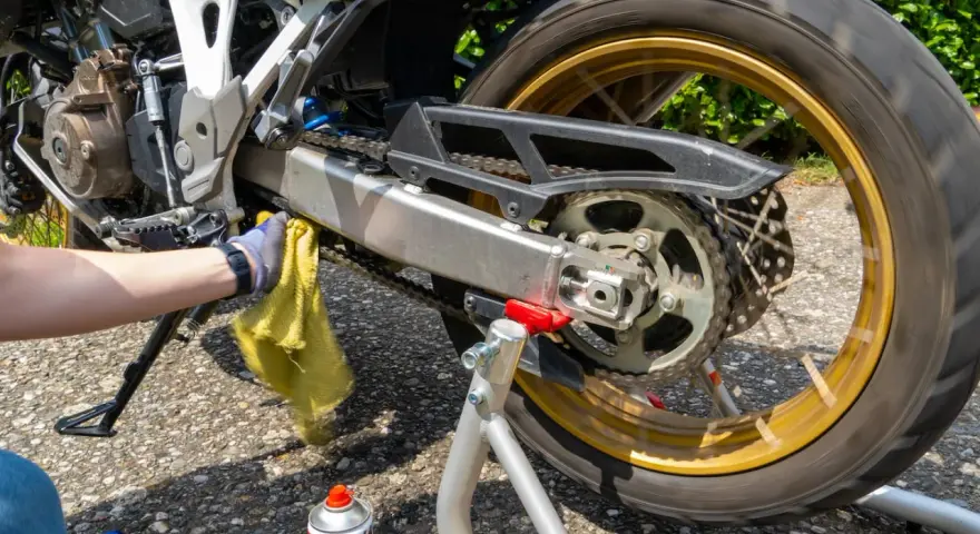 Mano guantata pulisce la catena della moto con uno straccio giallo. Spray per la pulizia catena visibile.