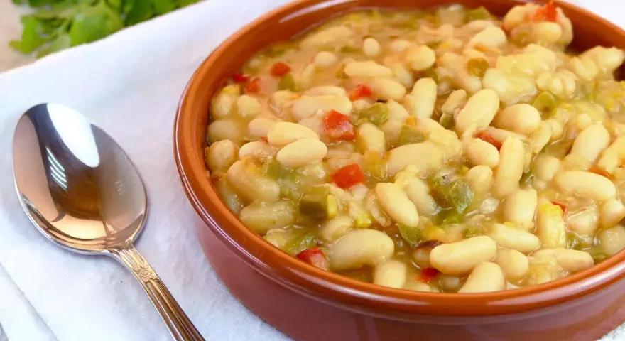 Un cuenco de barro lleno de alubias blancas con verduras, como pimientos rojos y verdes. Una cuchara plateada descansa a la izquierda.