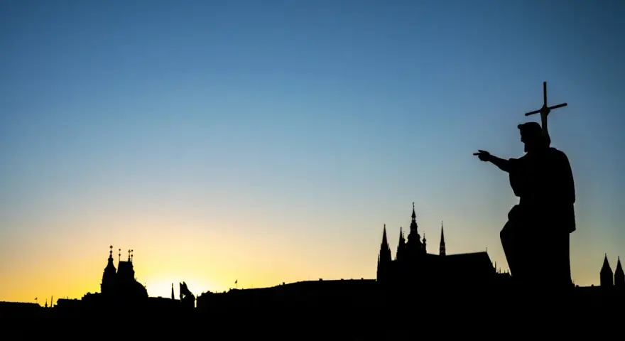 Silhouette d'une statue de saint pointant vers un paysage urbain au coucher du soleil. Le ciel bleu dégradé est le centre d'intérêt de l'image.