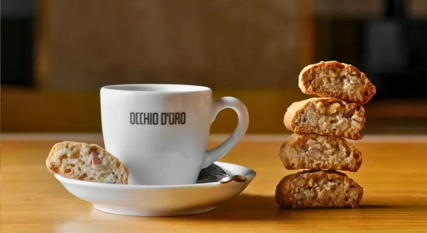Ein weißer Kaffeebecher mit "OCCHIO D'ORO" Schriftzug steht neben einem Stapel Cantuccini. Ein Cantuccino liegt auf der Untertasse. Dieses Bild inspiriert zum besten Cantuccini Rezept.
