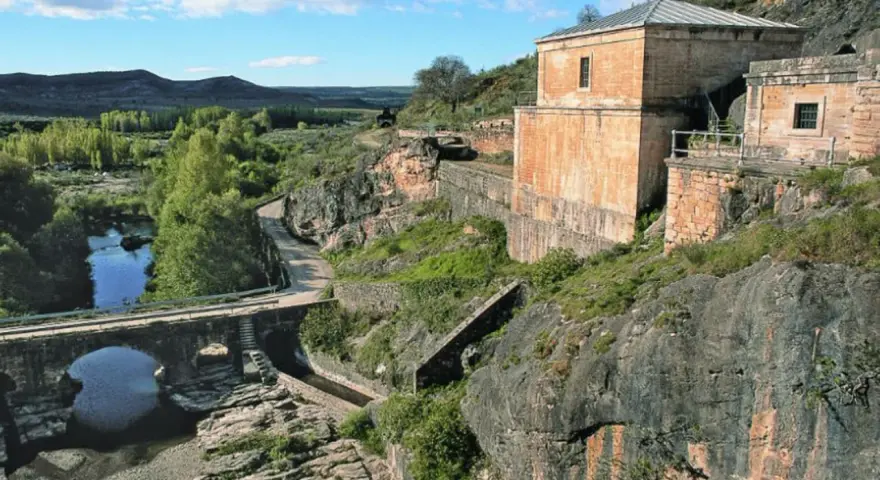 El portón de la oliva, un antiguo edificio de piedra junto a un río y un puente de arco, se alza sobre un paisaje rocoso y verde.