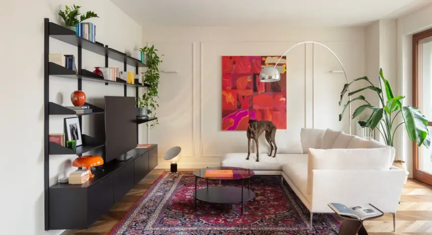 Un galgo se posa sobre un sofá blanco, rodeado de arte abstracto y un tapete persa. El **ruido visual** de los objetos decorativos y la pintura vibrante crea un ambiente ecléctico.