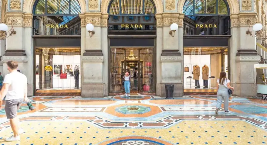Ingresso del negozio **Prada** con vetrine eleganti e un mosaico colorato sul pavimento. Persone passeggiano davanti alla boutique.