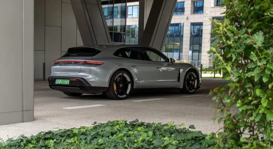 Szare Porsche Taycan Turbo S stoi pod zadaszeniem, z widokiem na nowoczesny budynek i zieleń.