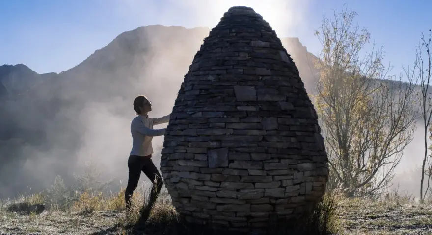 Une femme interagit avec une structure ovoïde en pierres, une œuvre de land art, dans un paysage brumeux et montagneux au lever du soleil.