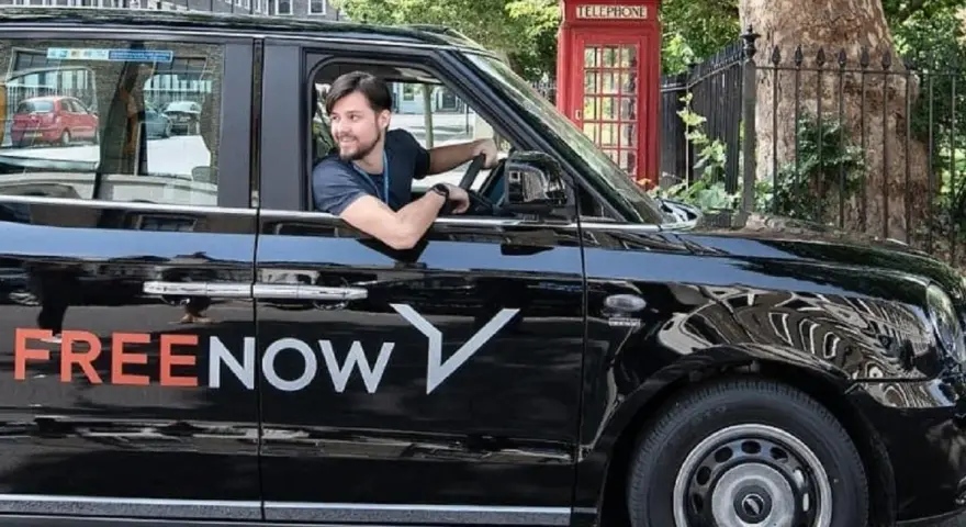Ein Mann fährt ein schwarzes freenow taxi. Im Hintergrund steht eine rote Telefonzelle.