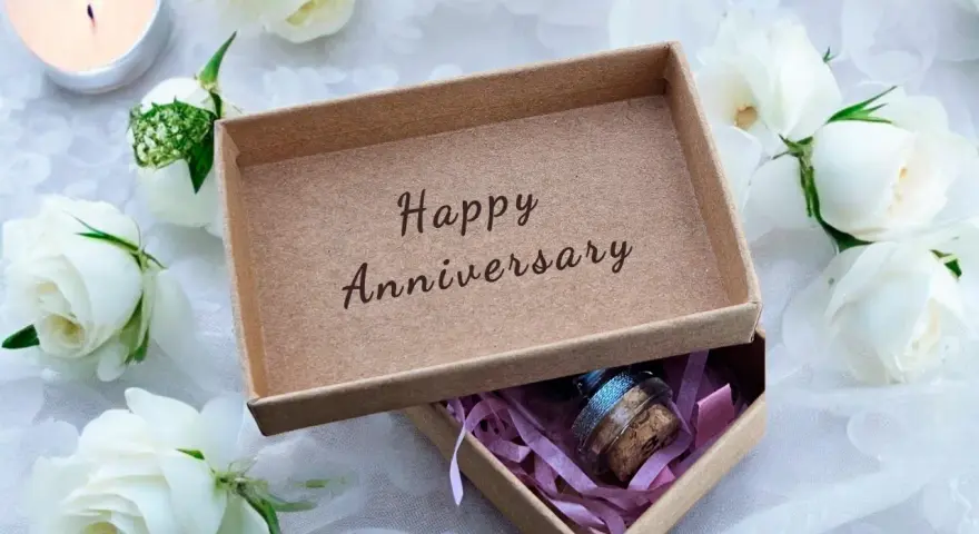 Otwarta szkatułka z napisem "Happy Anniversary" i małą buteleczką w środku, otoczona białymi różami i płonącą świecą. Idealny prezent na 21 rocznicę ślubu.