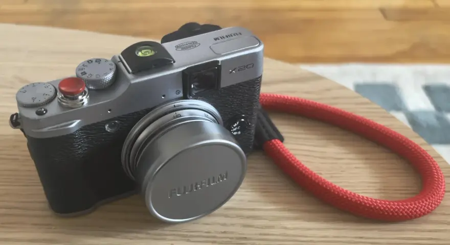 Srebrny aparat Fujifilm X20 z czerwoną smyczą leży na drewnianym blacie.