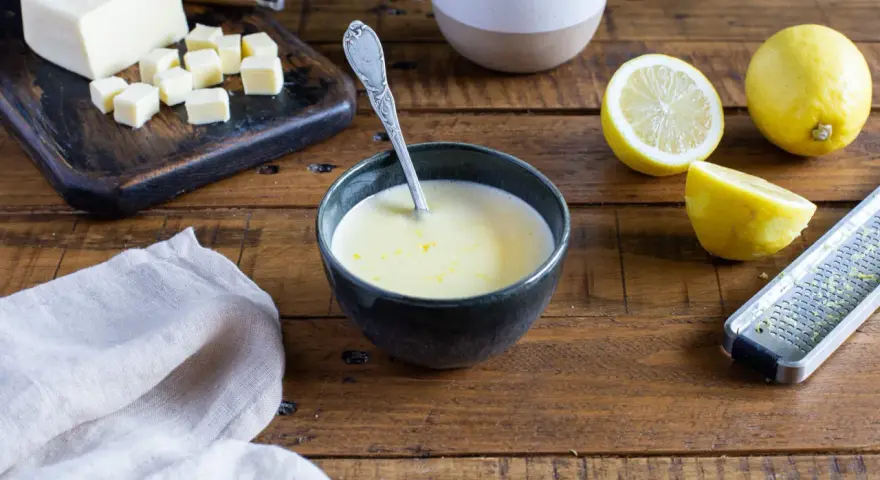 Un bol de sauce beurre citron, avec des citrons, du beurre coupé en dés et une râpe à zester sur une table en bois.