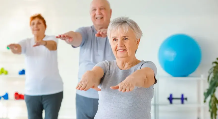 Trois seniors souriants pratiquent la gym douce, bras tendus, dans une salle lumineuse avec un ballon bleu en arrière-plan.