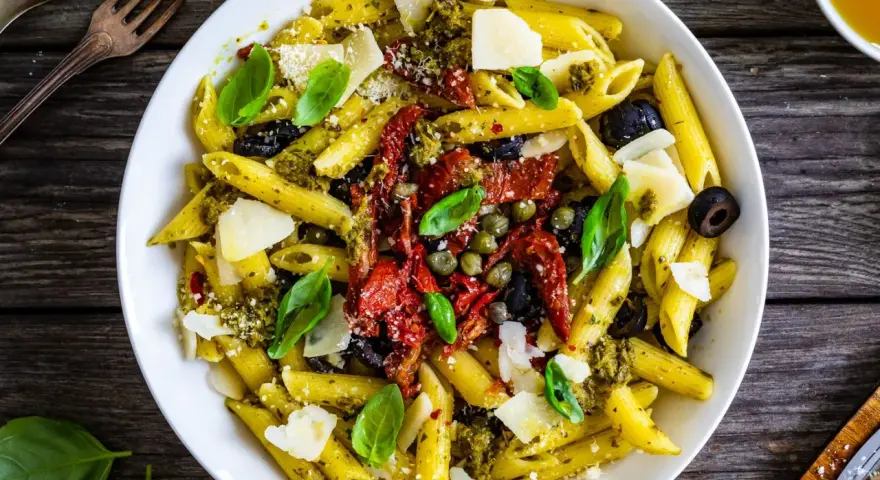 Penne con pesto, pomodori secchi, olive nere, capperi e scaglie di parmigiano. Un piatto colorato e saporito.